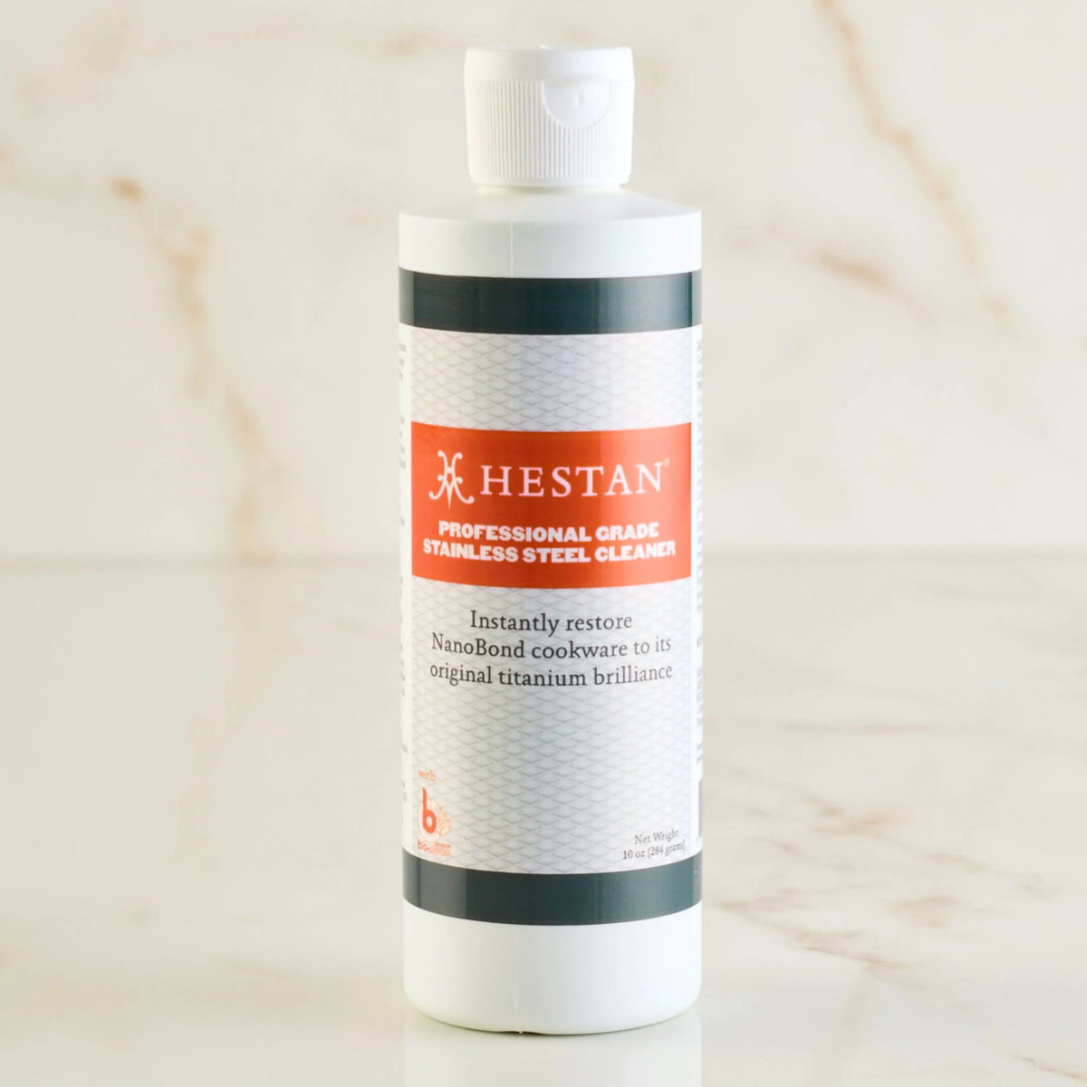 A white bottle of Accessories Hestan NanoBond® Professional Grade Cleaner with an orange label sits against a marble backdrop, showcasing its effectiveness in restoring the brilliance of NanoBond cookwares titanium surface, ensuring your kitchen shines like new.