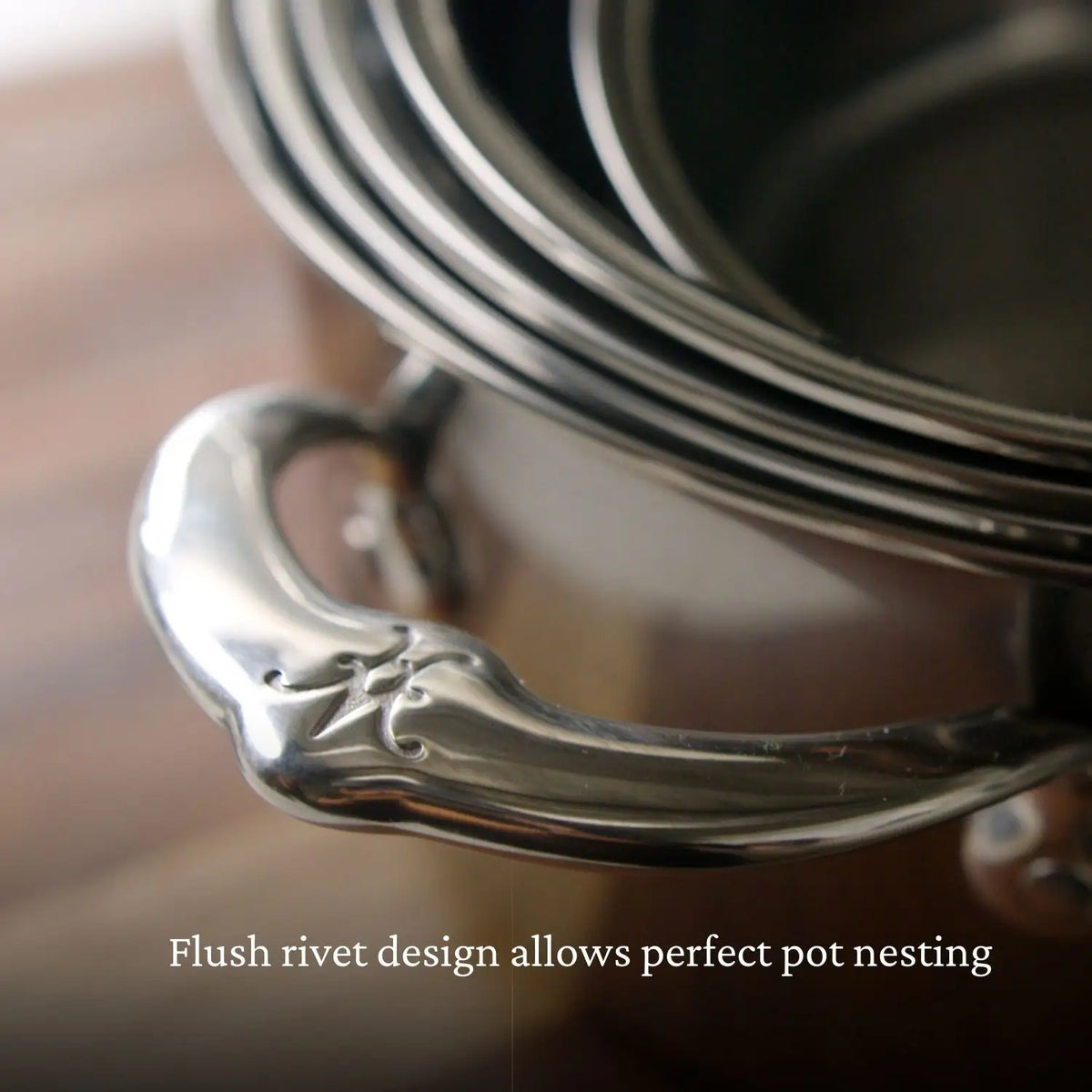 Close-up of nested stainless steel pots from the NanoBond® Titanium x Tyler Florence Cookware Set, 41-Piece, focusing on a decorative handle, with text overlay that reads, Flush rivet design allows perfect pot nesting.