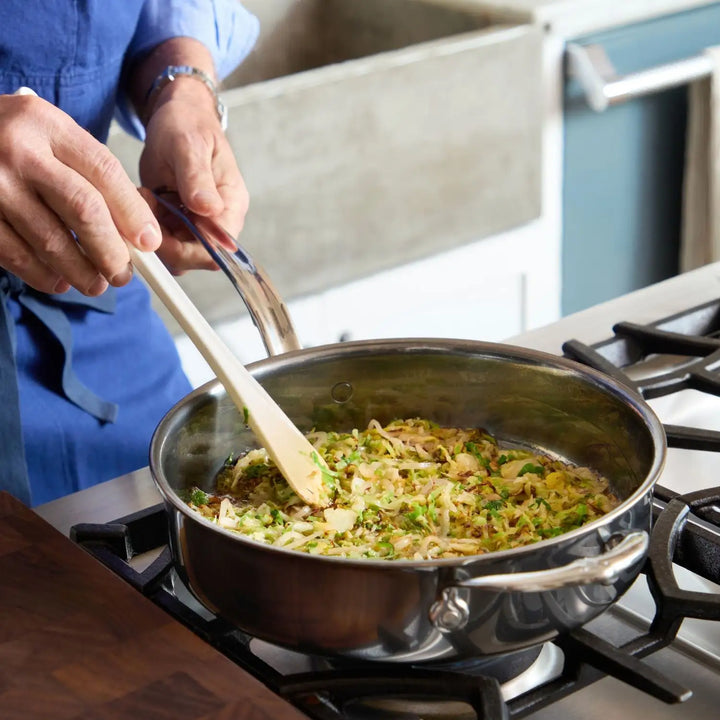 Wearing a blue shirt, someone uses a wooden spatula to stir a dish in the NanoBond 10-Piece Titanium Precision Saucepan Cookware Set on a gas stove. The stainless steel saucepan glistens while cooking vibrant vegetables like sliced onions and chopped green herbs.