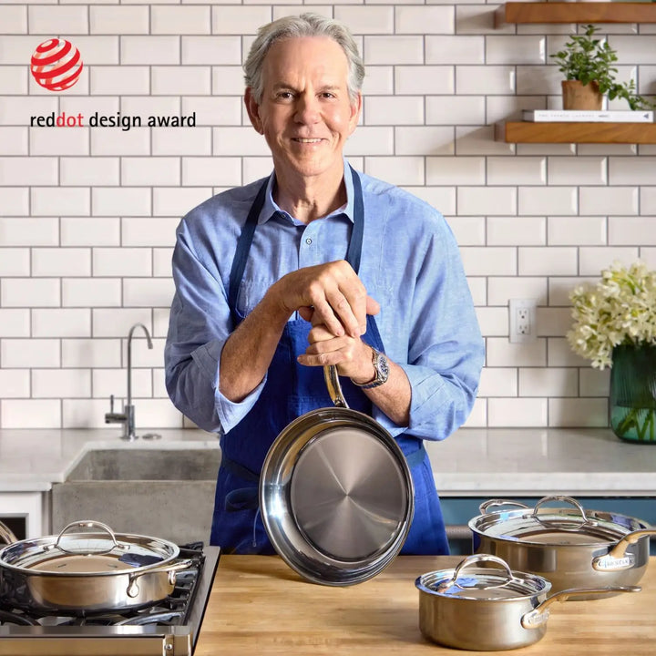 In a kitchen adorned with a Red Dot Design Award logo, a person in a blue apron holds a saucepan from the NanoBond 10-Piece Titanium Precision Cookware Set. Pots scatter across the counter, while white subway tiles and wooden shelves with plants complete the scene, ensuring stainless steel cookware shines.