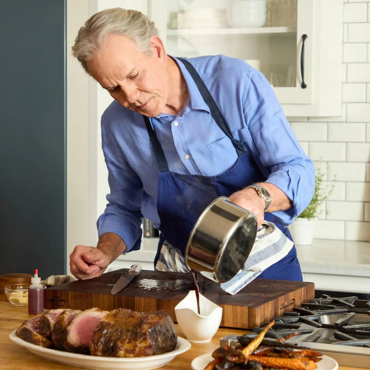 Wearing a blue shirt and apron, an older man expertly pours sauce from a NanoBond 10-Piece Titanium Precision Saucepan onto a cutting board in his kitchen. A plate of sliced meat and vegetables is on the counter, framed by white tiles and sleek cabinets.