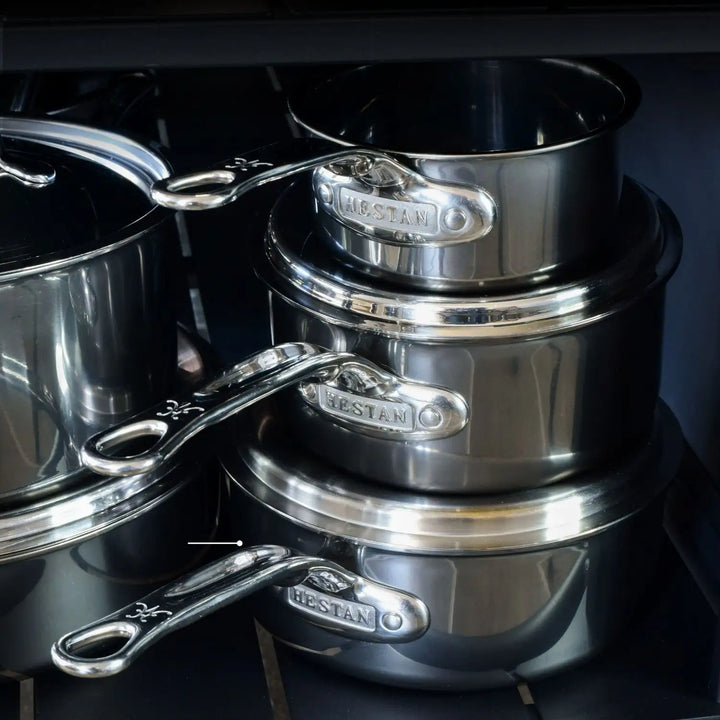 Three shiny saucepans with handles, part of the 10-Piece Titanium Precision Saucepan Cookware Set by NanoBond, are neatly stacked on a shelf. Additional stainless steel cookware can be seen to the left in what looks like a kitchen or cookware display area.