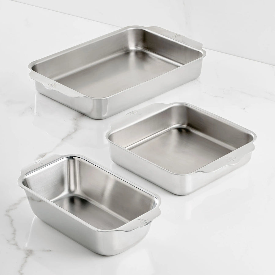 The Hestan Provisions OvenBond Tri-Ply 3-Piece Baking Set, featuring three stainless steel pans with rounded corners and side handles for easy handling, is displayed on a white marble surface.