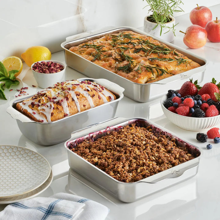 On the kitchen counter, three baked dishes rest: a rectangular bread with icing and flowers, a square crumble, and rosemary focaccia. Nearby are fruit and dishware. All were prepared using the Hestan Provisions OvenBond Tri-Ply 3-Piece Baking Set for consistent results.