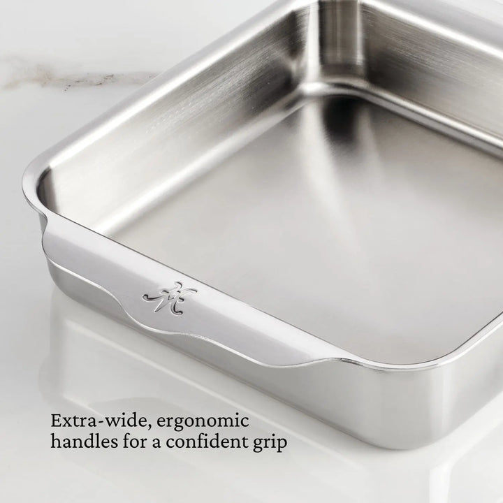 Close-up of an OvenBond Tri-Ply baking pan from Hestan Provisions on a marble surface. The pan has ergonomic extra-wide handles for a firm grip. Text reads, Extra-wide, ergonomic handles for confident grip and warp-resistant durability.