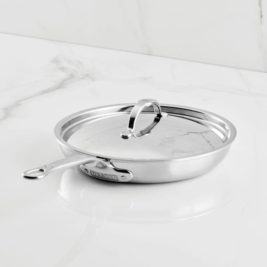 A ProBond® Luxe™ Polished Clad Stainless Steel Covered Skillet with a matching lid sits on a white marble countertop, featuring metal handles and a polished, reflective finish.