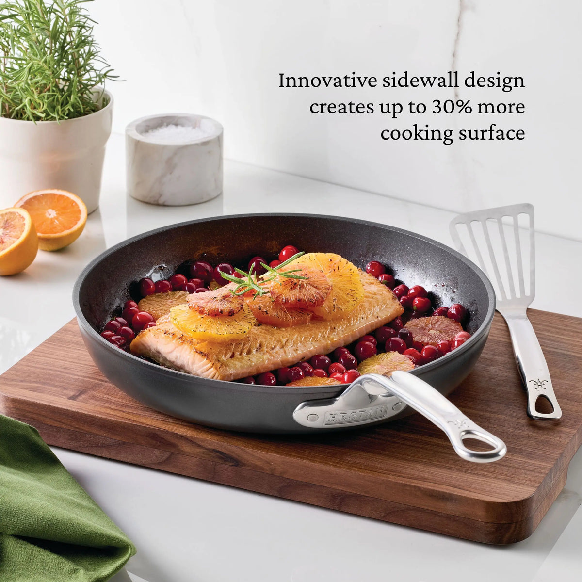 A cooked salmon fillet with orange slices and herbs sits in a TITUM® Fusion TITUM Nonstick Cookware Set skillet, induction compatible, with cranberries. The skillet is on a wooden board beside a spatula, sliced orange, marble container, and rosemary plant. Text is overlaid.