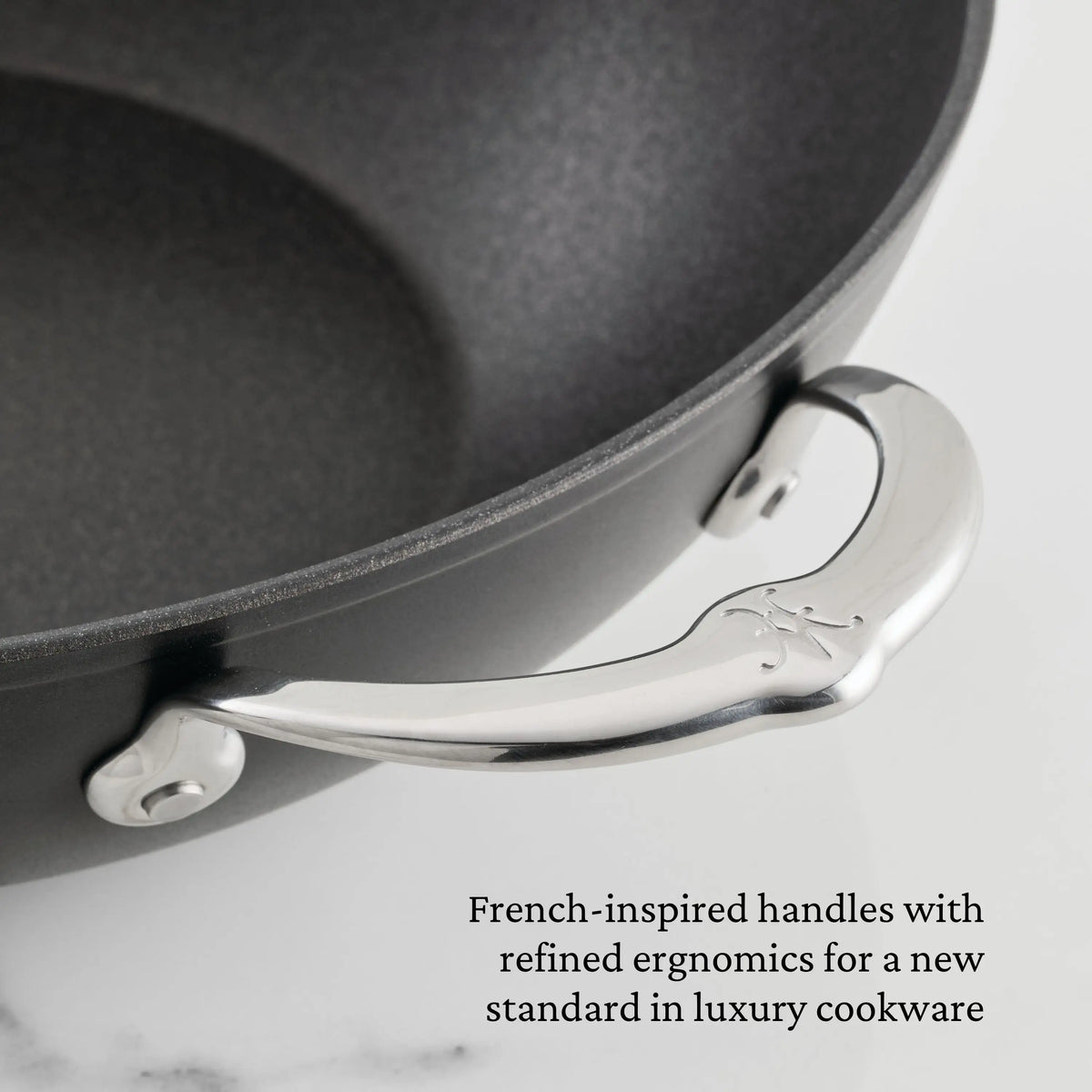 Close-up of a TITUM® Fusion TITUM Nonstick Cookware Set pan featuring a curved stainless steel handle with two rivets. Text notes French-inspired, ergonomic handles handcrafted in Italy for this luxury 10-piece nonstick set.