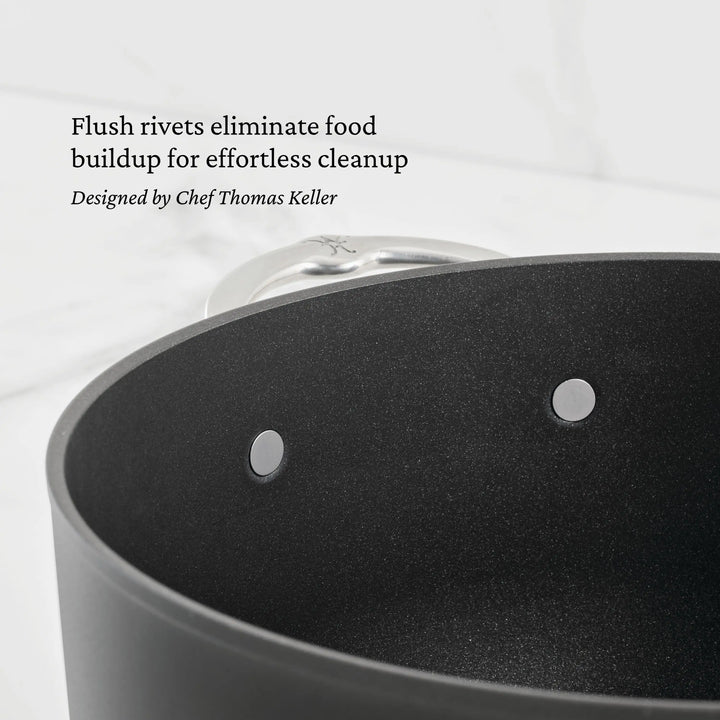 A close-up of a black TITUM® Fusion pot shows two flush rivets inside. Text above reads, Flush rivets eliminate food buildup for easy cleanup. This TITUM Nonstick Cookware Set, 10-Piece, is designed by Chef Thomas Keller.