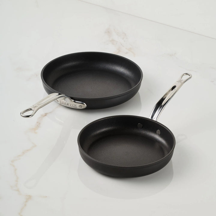 Two black TITUM® Fusion TITUM Nonstick Open Skillets with metal handles rest on a white marble surface; handcrafted in Italy, the larger has a straight handle and the smaller has a gently curved one.