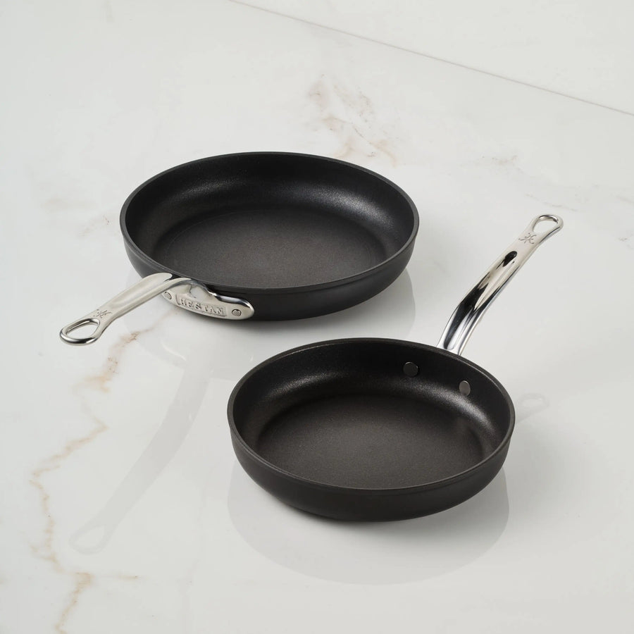 Two black TITUM® Fusion TITUM Nonstick Open Skillets with metal handles rest on a white marble surface; handcrafted in Italy, the larger has a straight handle and the smaller has a gently curved one.