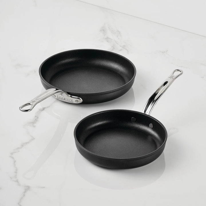 Two black TITUM Fusion™ Nonstick Open Skillets with silver handles are placed on a white marble surface. Highlighting their durable quality, the larger skillet is positioned above and to the left of the smaller one.