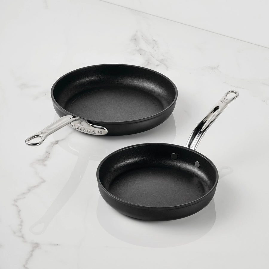 Two black TITUM Fusion™ Nonstick Open Skillets with silver handles are placed on a white marble surface. Highlighting their durable quality, the larger skillet is positioned above and to the left of the smaller one.