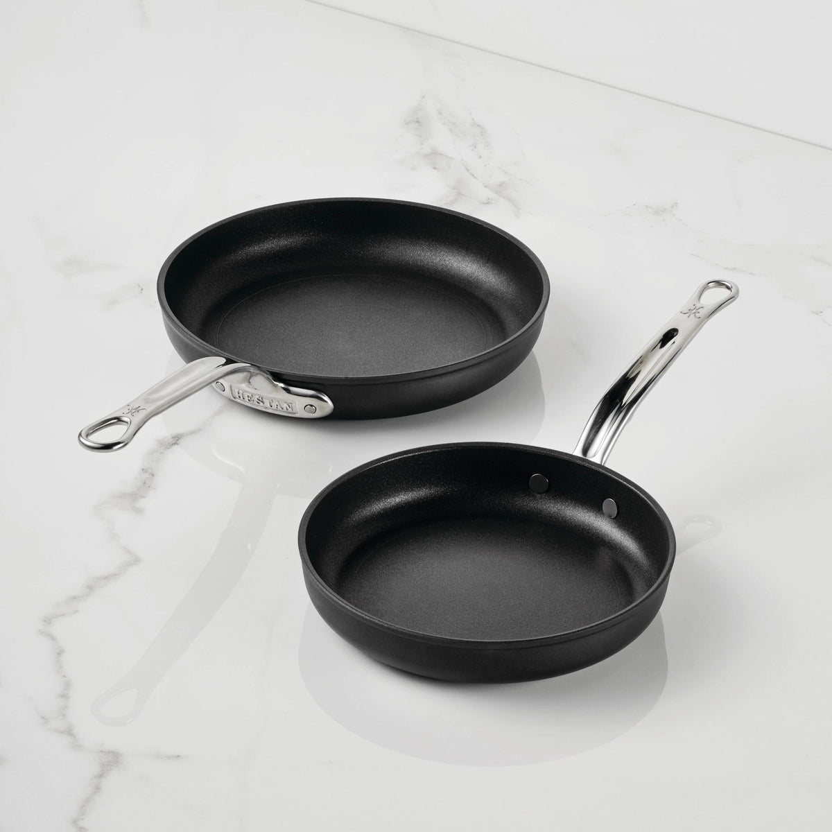 Two black TITUM Fusion™ Nonstick Open Skillets with silver handles are placed on a white marble surface. Highlighting their durable quality, the larger skillet is positioned above and to the left of the smaller one.