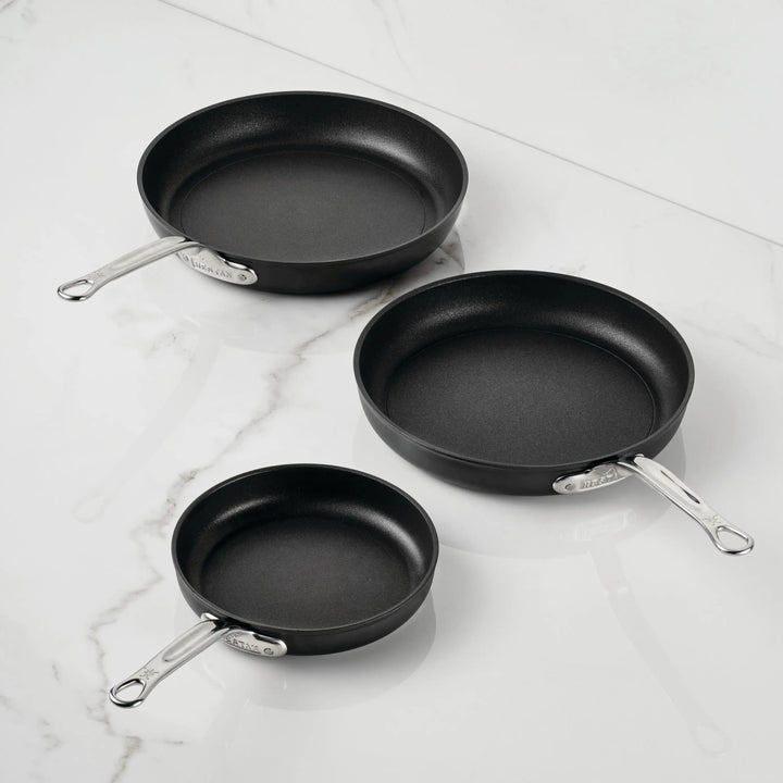 Three TITUM® Fusion TITUM Nonstick Open Skillets in varying sizes, featuring silver handles, are arranged on a white marble surface with each handle pointing a different direction.