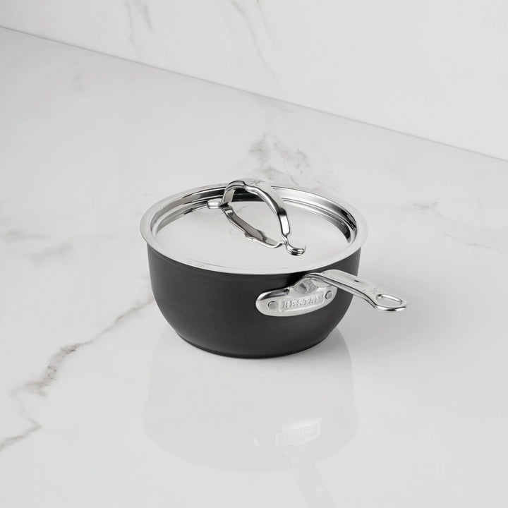 A small black TITUM® Fusion TITUM Nonstick Saucepan with a stainless steel handle and lid sits on a white marble countertop. Induction compatible, it features a metal label displaying the brand on its side.