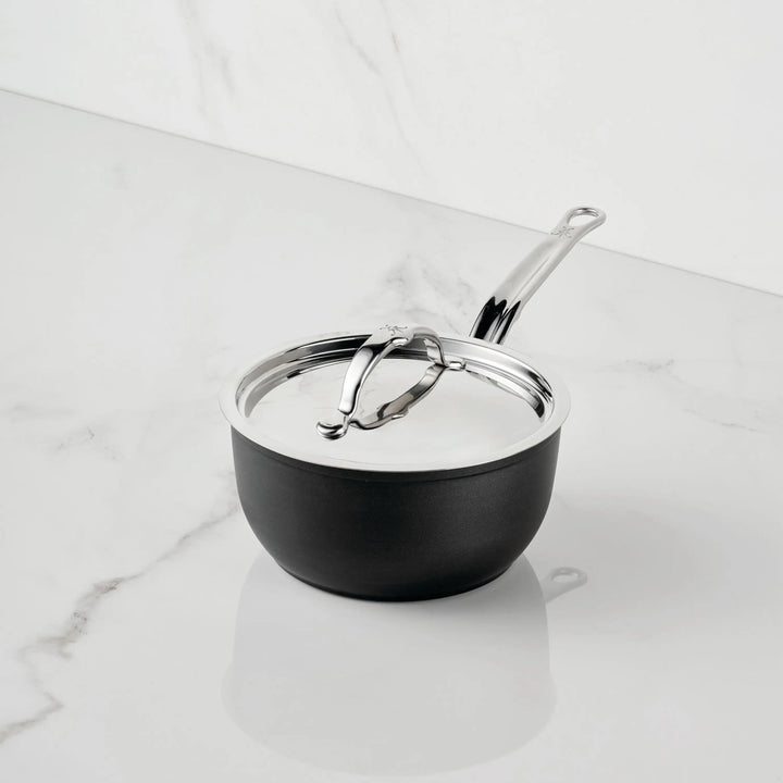The TITUM® Fusion TITUM Nonstick Saucepans with Lids, featuring a sleek black design, metal lid, and long silver handle, sits on a white marble countertop. Induction compatible, it combines style and versatility for modern kitchens.