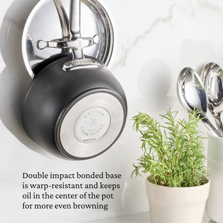 A TITUM® Fusion TITUM Nonstick Saucepan hangs on a metal hook above a potted herb. Two metal spoons rest nearby. Text notes the saucepan’s warp-resistant, even-heating base. Handcrafted in Italy.