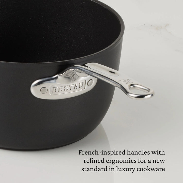A close-up of a black TITUM® Fusion saucepan with a silver handle engraved with HEYSTAN. Text reads: French-inspired ergonomic handles highlight Italian-crafted excellence in luxury TITUM Nonstick Saucepans with Lids.