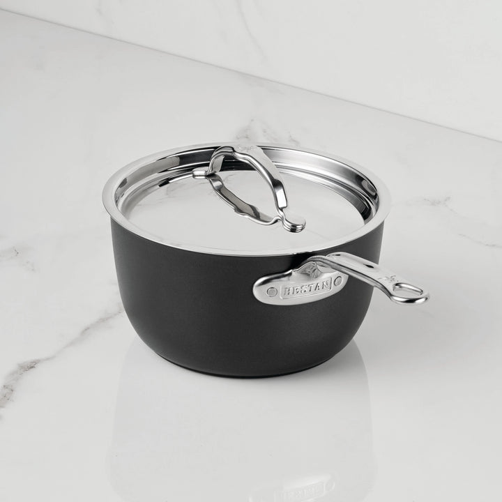 A black and silver saucepan with a lid from the TITUM Fusion™ TITUM Nonstick Saucepans with Lids collection sits on a white marble countertop. Featuring a metal handle and a label that reads LEGACY, this pan’s double impact stainless steel base ensures lasting durability and even heat distribution. The background is a plain white wall.
