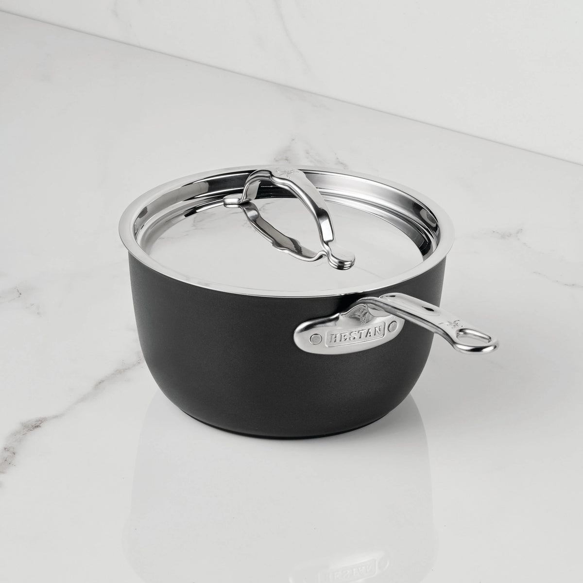 A black and silver saucepan with a lid from the TITUM Fusion™ TITUM Nonstick Saucepans with Lids collection sits on a white marble countertop. Featuring a metal handle and a label that reads LEGACY, this pan’s double impact stainless steel base ensures lasting durability and even heat distribution. The background is a plain white wall.