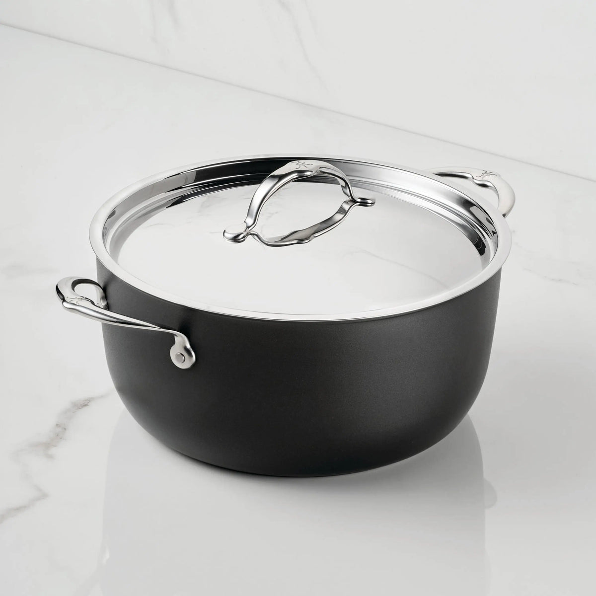 The TITUM® Fusion TITUM Nonstick Stock Pot with Lid, 8-Quart, features a black round design with two metal handles and a shiny silver lid, handcrafted in Italy and shown on a white marble surface against a marble background.