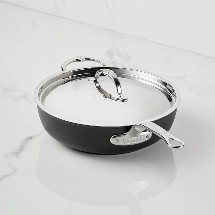 A black and silver TITUM® Fusion Nonstick Essential Pan with Lid, 4.5-Quart, rests on a white marble countertop. Both the metallic handle and glass lid feature the brand’s nameplate, showcasing this induction-ready cookware.
