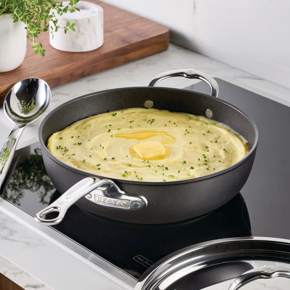 A pan of creamy mashed potatoes with chives and butter sits in the TITUM® Fusion TITUM Nonstick Essential Pan with Lid, 4.5-Quart, on a stovetop. A spoon and fresh herbs rest nearby on a white marble countertop.