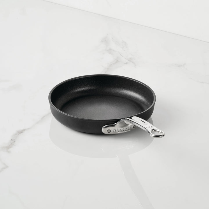 A small black TITUM Nonstick Open Skillet from TITUM Fusion™ with a silver handle sits on a white marble countertop. The handle displays the TITUM Fusion™ brand name, emphasizing the high quality of this induction-compatible nonstick cookware. The skillet is empty and spotless.