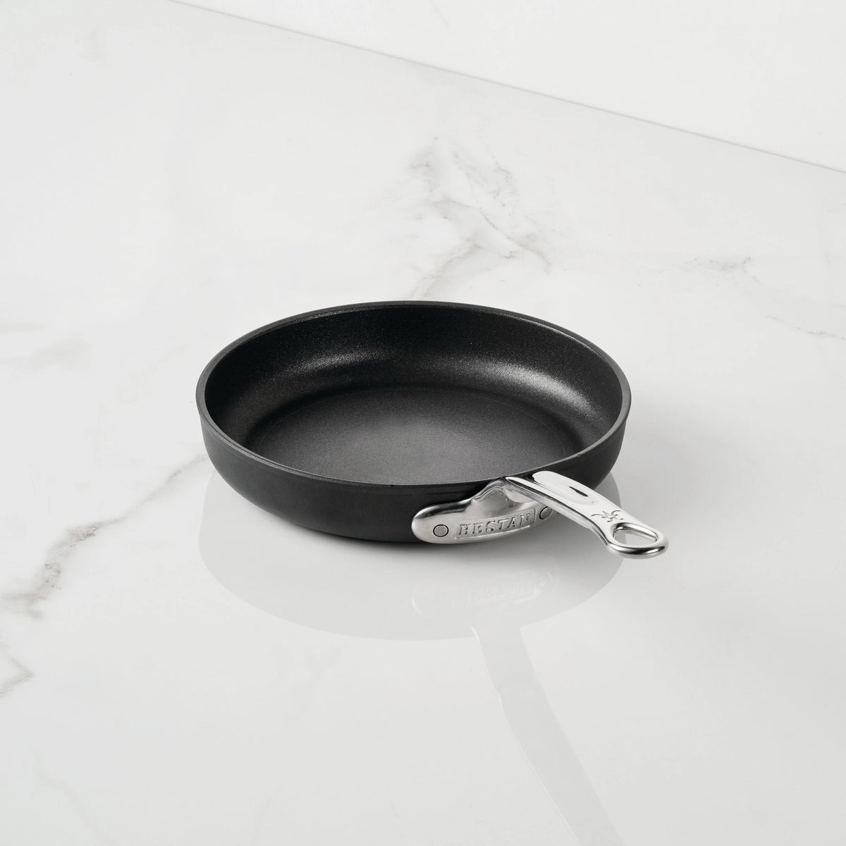 A small black TITUM Nonstick Open Skillet from TITUM Fusion™ with a silver handle sits on a white marble countertop. The handle displays the TITUM Fusion™ brand name, emphasizing the high quality of this induction-compatible nonstick cookware. The skillet is empty and spotless.