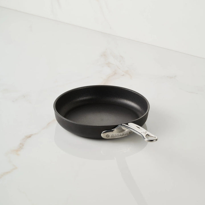 A clean, empty TITUM® Fusion TITUM Nonstick Open Skillet with a metal handle rests on a white marble surface. Handcrafted in Italy, the small black pan stands out with no utensils or food around it.