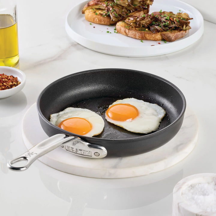 The TITUM® Fusion TITUM Nonstick Open Skillets, handcrafted in Italy, holds two sunny-side-up eggs on a marble surface with toast, olive oil, red pepper flakes, and salt in the background.