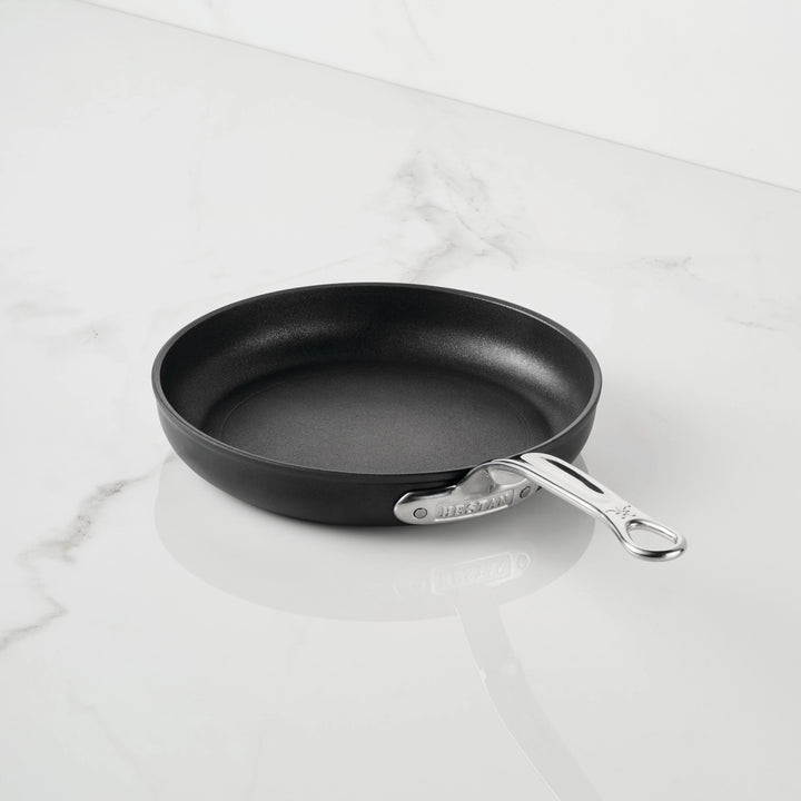 The TITUM Fusion™ TITUM Nonstick Open Skillets, part of premium nonstick cookware, features a black nonstick surface and a sleek silver metal handle. Resting empty at a slight angle on a white marble surface with its handle pointing to the right, this durable skillet combines modern design with lasting performance.
