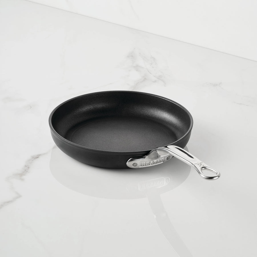 The TITUM Fusion™ TITUM Nonstick Open Skillets, part of premium nonstick cookware, features a black nonstick surface and a sleek silver metal handle. Resting empty at a slight angle on a white marble surface with its handle pointing to the right, this durable skillet combines modern design with lasting performance.