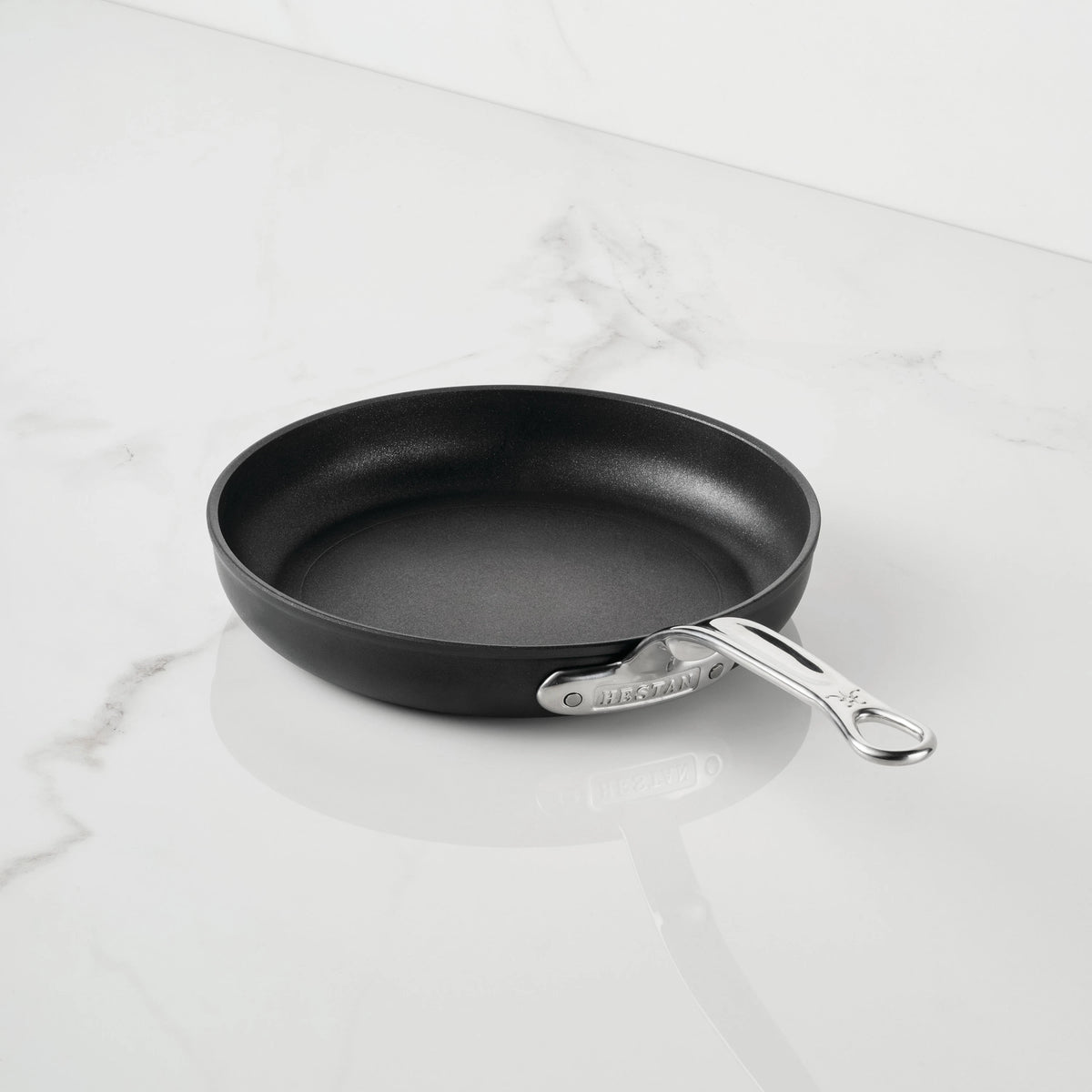 The TITUM Fusion™ TITUM Nonstick Open Skillets, part of premium nonstick cookware, features a black nonstick surface and a sleek silver metal handle. Resting empty at a slight angle on a white marble surface with its handle pointing to the right, this durable skillet combines modern design with lasting performance.