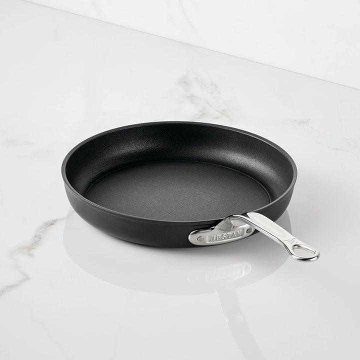 A TITUM Fusion™ TITUM Nonstick Open Skillet with a sleek metal handle rests on a white marble countertop, showcasing the elegance of durable Italian craftsmanship.
