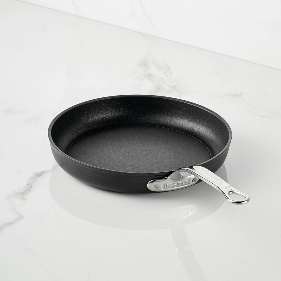 A TITUM Fusion™ TITUM Nonstick Open Skillet with a sleek metal handle rests on a white marble countertop, showcasing the elegance of durable Italian craftsmanship.