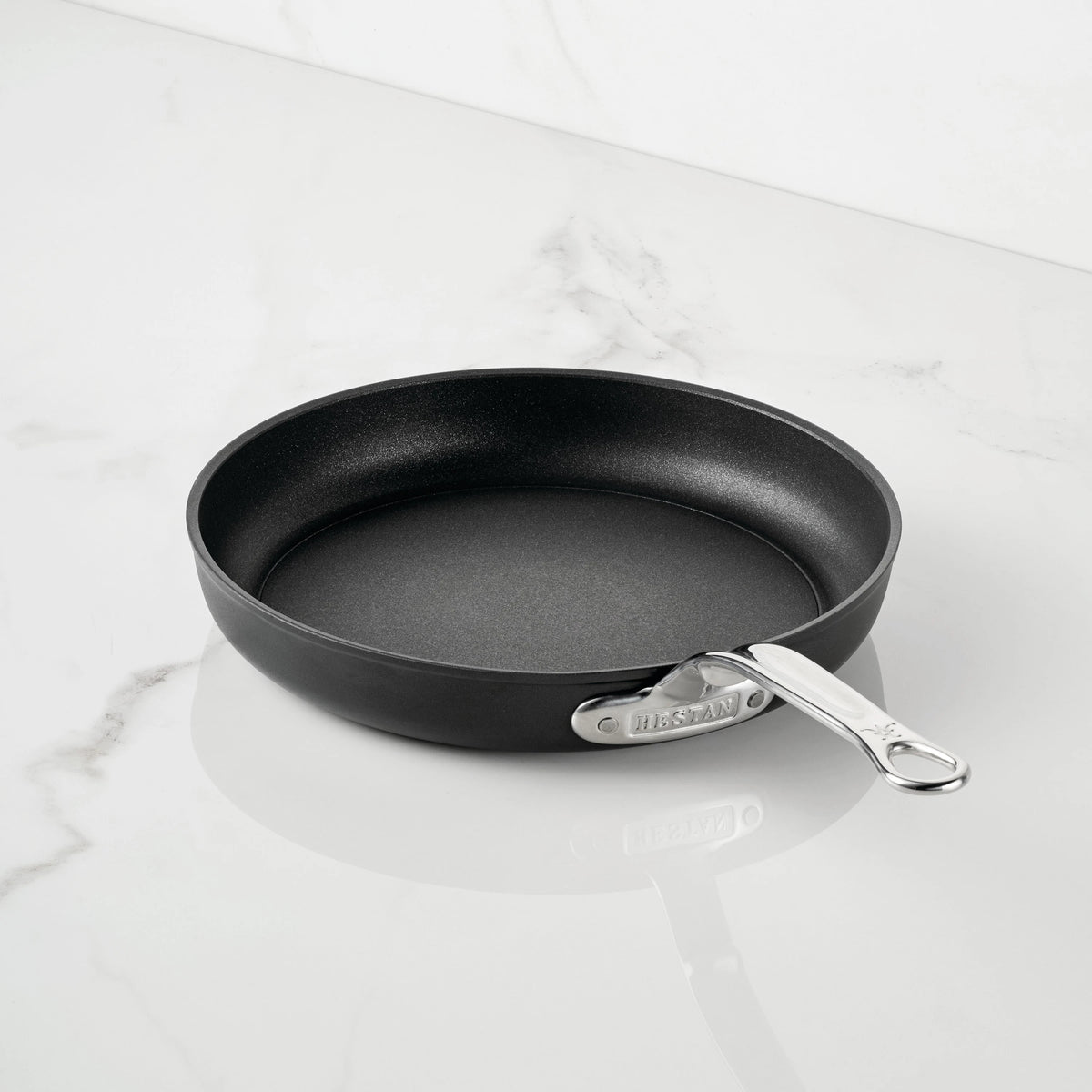 A TITUM Fusion™ TITUM Nonstick Open Skillet with a sleek metal handle rests on a white marble countertop, showcasing the elegance of durable Italian craftsmanship.