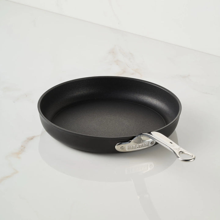 A TITUM® Fusion TITUM Nonstick Open Skillet with a metal handle, handcrafted in Italy, sits empty on a white marble countertop. The sleek black pan is shown from above at a slight angle.