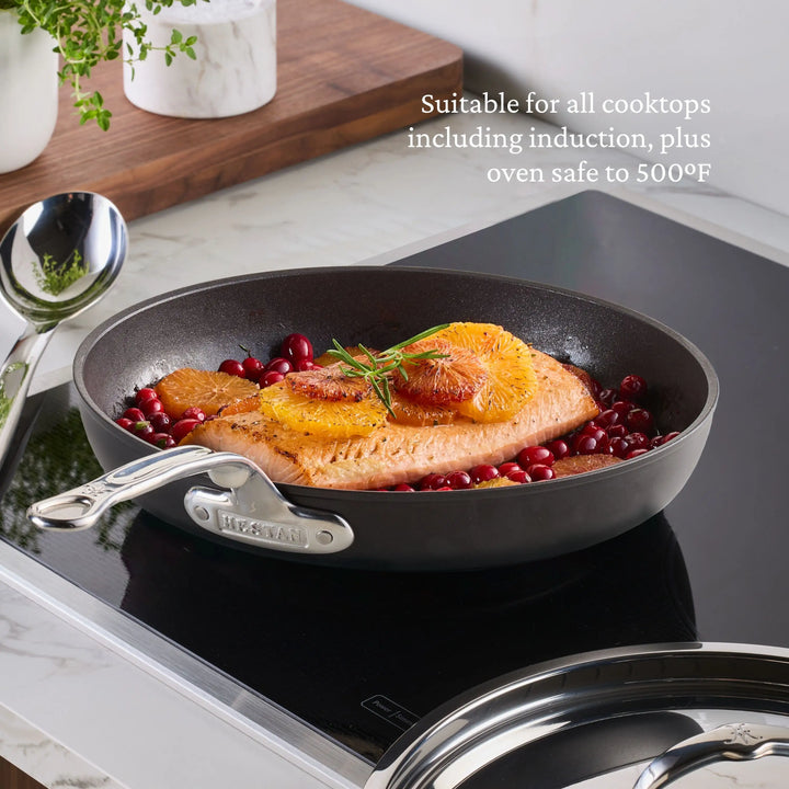 A salmon fillet with oranges and herbs cooks with cranberries in the TITUM® Fusion 11-Inch TITUM Nonstick Skillet with Lid on an induction cooktop. White text notes it's handcrafted in Italy, oven safe to 500°F, and works on all cooktops.