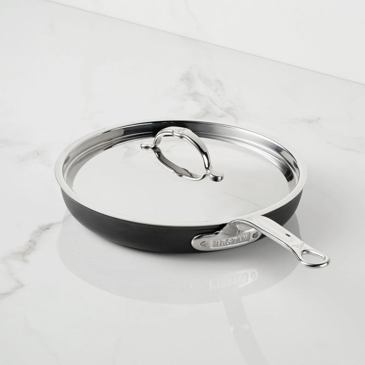 An 11-inch TITUM® Fusion Nonstick Skillet with Lid in black and stainless steel rests on a white marble surface, featuring a long silver handle engraved with the brand name.