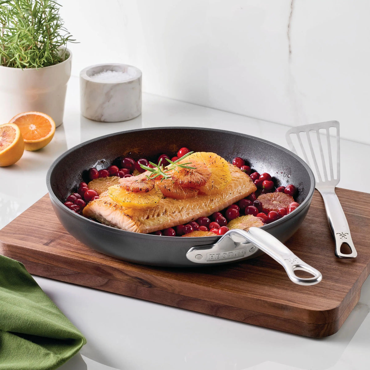 A cooked salmon fillet with orange slices and rosemary is served in the TITUM® Fusion TITUM Nonstick Skillet with Lid, 11-Inch, resting on a wooden board amid cranberries, citrus, a spatula, green napkin, and cut citrus on a white countertop.