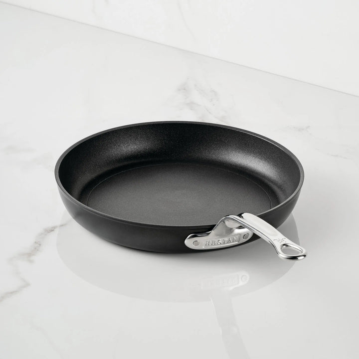 A TITUM® Fusion TITUM Nonstick Open Skillet with a silver riveted handle, handcrafted in Italy, rests empty on a white marble countertop—an elegant example of Italian nonstick cookware.