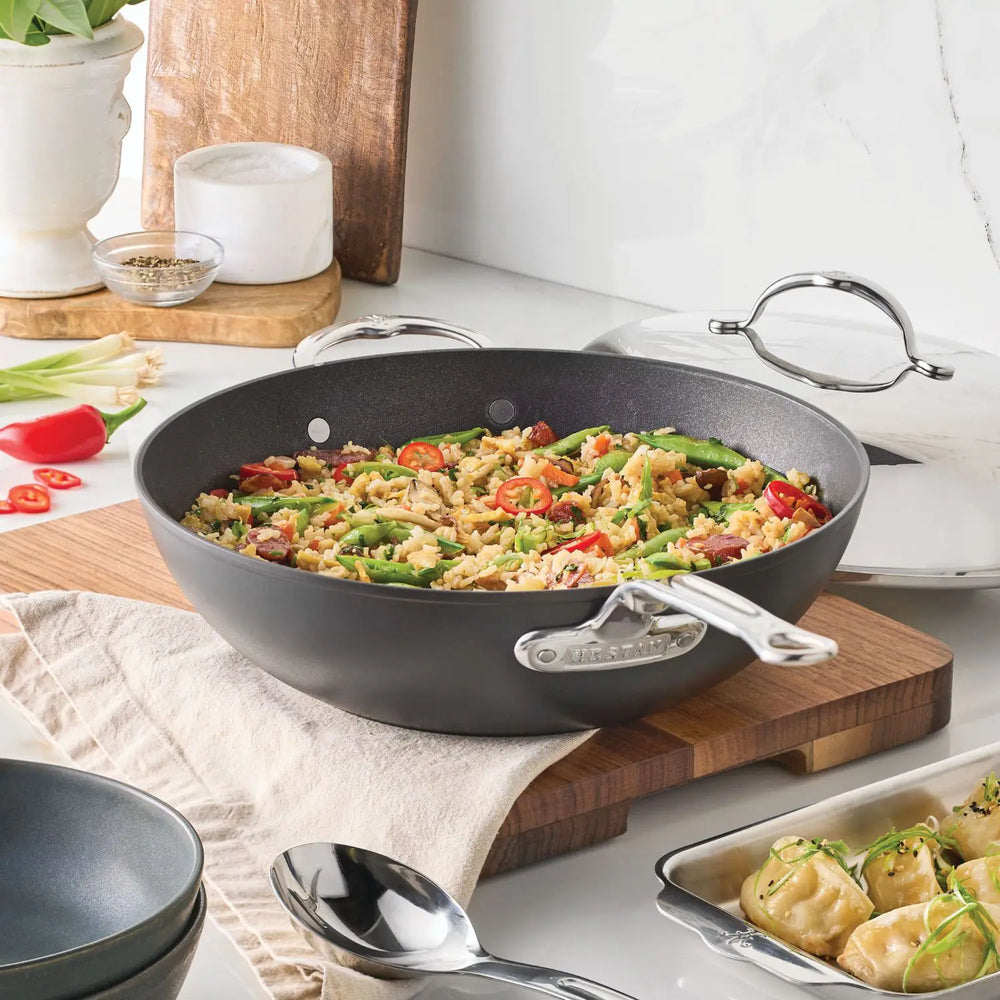 The TITUM® Fusion TITUM Nonstick Wok with Stick Handle and Dome Lid, 12.5-Inch, sits on a cutting board filled with stir-fried rice and vegetables. Nearby are a serving spoon, a bowl, and dumplings on a plate garnished with greens.