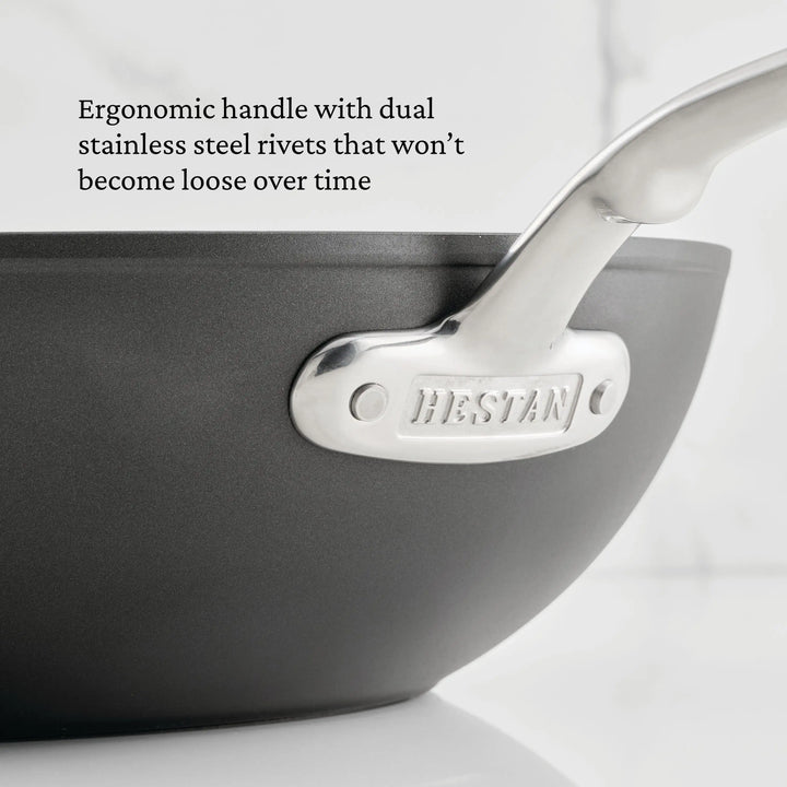 Close-up of a 12.5-inch TITUM® Fusion Nonstick Wok with Stick Handle and Dome Lid; metal handle secured by two stainless steel rivets, “TITUM” engraved on the bracket. Text highlights its ergonomic design and durable nonstick surface. Handcrafted in Italy.