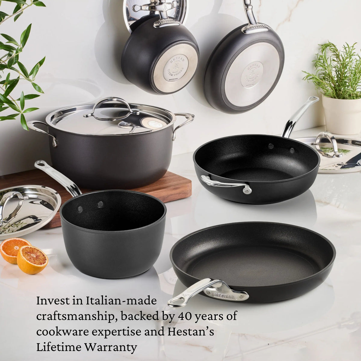 The TITUM® Fusion 12.5-Inch Nonstick Wok with Stick Handle and Dome Lid, handcrafted in Italy, sits on a white countertop with oranges and greenery. Three pans hang behind. Text highlights Hestan’s warranty.