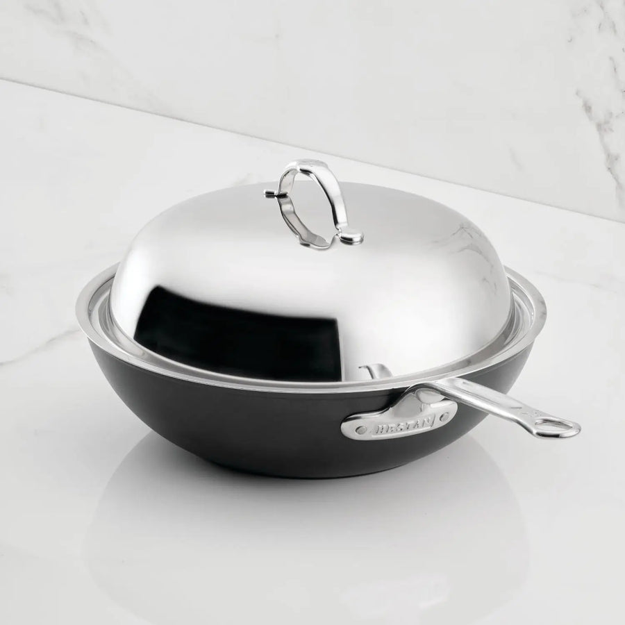 A black and silver TITUM® Fusion 12.5-Inch Nonstick Wok with stick handle and polished dome lid sits on a white marble surface, highlighting refined Italian craftsmanship.