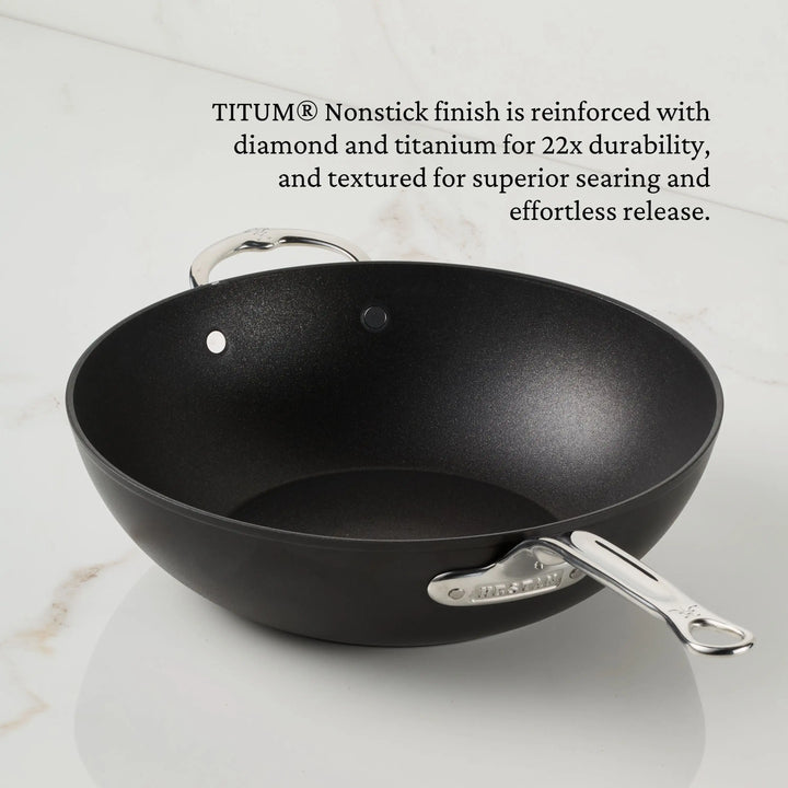 A black TITUM® Fusion 12.5-Inch Nonstick Wok with a stick handle and dome lid sits on a white surface. Its durable nonstick finish, handcrafted in Italy, is reinforced with diamond and titanium for long-lasting performance.