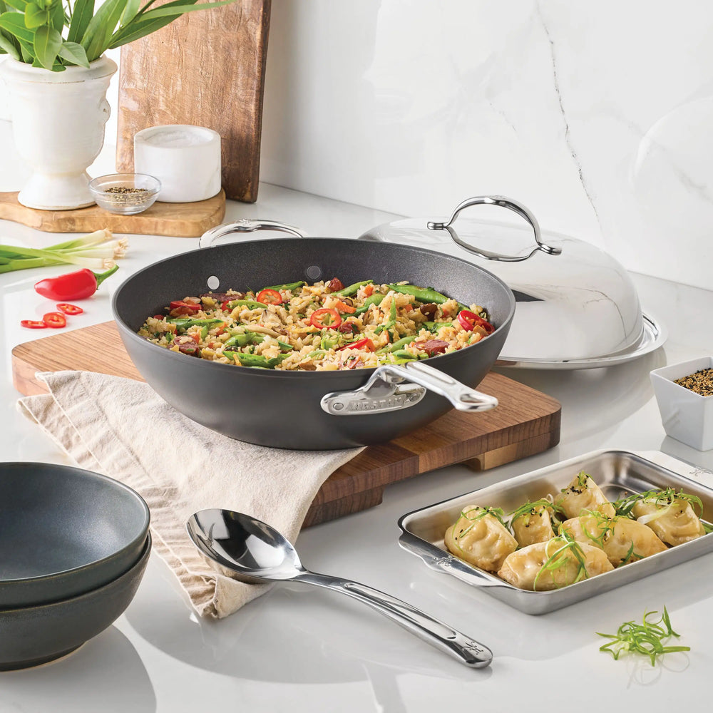 A TITUM® Fusion 12.5-Inch TITUM Nonstick Wok with Stick Handle and Dome Lid filled with fried rice and vegetables sits on a wooden board, surrounded by a serving spoon, stacked bowls, dumplings on a tray, and chopped green onions on a white counter.