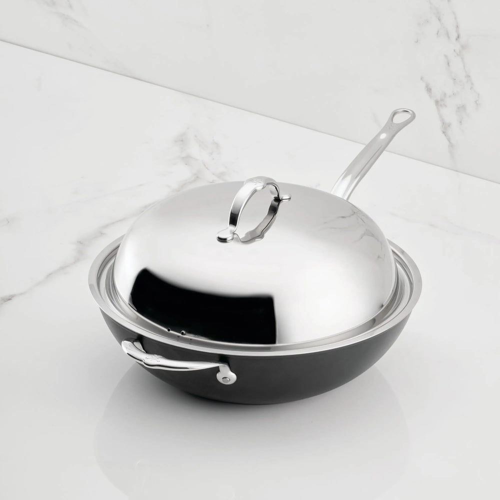 The TITUM® Fusion 12.5-Inch Nonstick Wok with Stick Handle and Dome Lid features a matte black finish, double impact stainless steel, and a side handle. Handcrafted in Italy, it sits on a white marble surface.